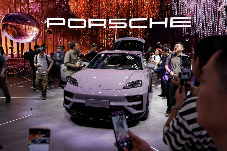 A imagem mostra um carro da marca Porsche em um evento， cercado por várias pessoas que observam e tiram fotos. O carro é de cor clara e está em destaque， com um fundo que apresenta uma iluminação vibrante e elementos decorativos. Acima do carro， há um grande letreiro com a palavra 039;PORSCHE039;.