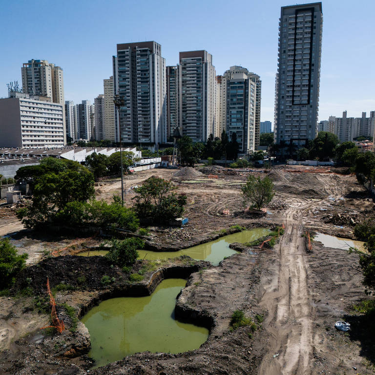 A imagem mostra uma área em construção com terrenos desmatados e algumas poças de água verde. Ao fundo, há vários edifícios altos e modernos. O céu está claro e ensolarado, e a vegetação é visível em algumas partes do terreno.