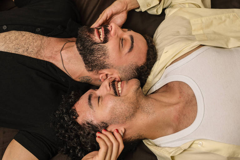 A imagem mostra dois homens deitados lado a lado， sorrindo e se olhando. Um deles tem cabelo encaracolado e barba， enquanto o outro tem cabelo liso e uma leve barba. Eles estão em um ambiente aconchegante， com roupas casuais. Um dos homens está usando uma camiseta preta e o outro uma camiseta branca， com uma camisa amarela aberta por cima.