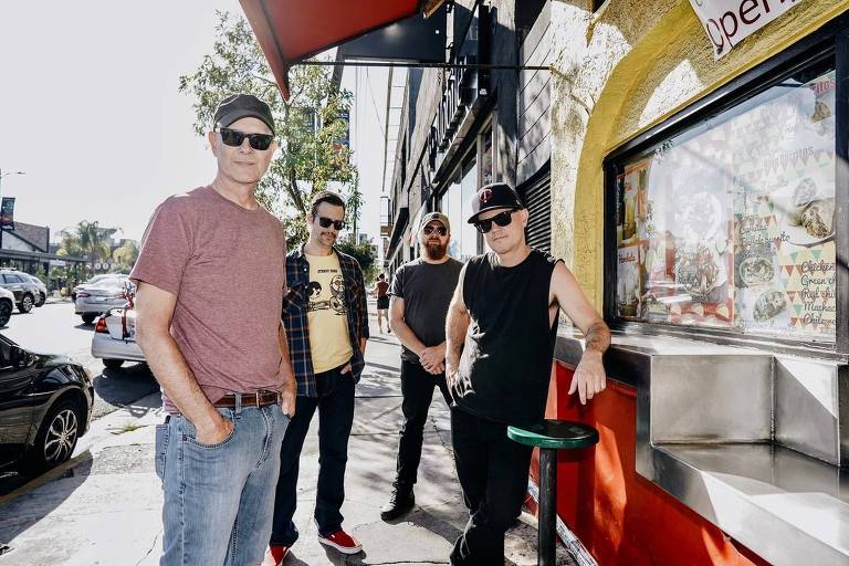 Quatro homens estão posando em frente a um restaurante. O primeiro à esquerda usa uma camiseta rosa e jeans, o segundo usa uma camiseta amarela com uma estampa e uma camisa xadrez por cima, o terceiro tem uma barba e usa uma camiseta cinza, e o último está usando uma camiseta preta sem mangas e um boné. Ao fundo, há um cardápio visível na janela do restaurante, e a rua está movimentada com carros e pessoas.