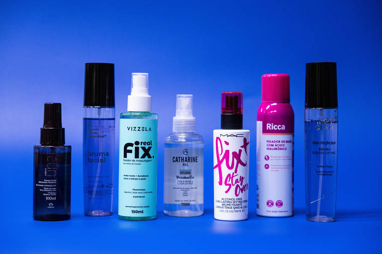 A imagem apresenta uma linha de produtos de beleza dispostos em uma superfície azul. Os frascos variam em tamanho e formato， com rótulos visíveis. Os produtos incluem sprays e finalizadores， com marcas como Vizzela， Catharine， MAC e Ricca. Os frascos têm cores variadas， incluindo preto， azul， rosa e transparente.