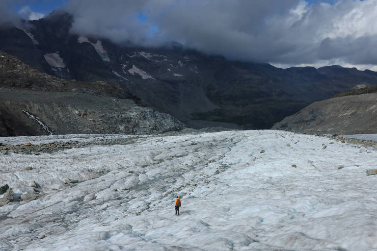 A imagem mostra um explorador solitário caminhando sobre um grande glaciar. O cenário é montanhoso， com nuvens escuras no céu e uma paisagem de gelo e rochas ao redor. O explorador está vestido com uma jaqueta laranja， destacando-se contra o fundo gelado.