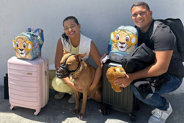 A imagem mostra um casal posando ao lado de duas malas coloridas com estampas de animais. A mulher está vestindo uma blusa branca e uma saia clara， enquanto o homem usa uma camiseta preta e jeans. Eles estão agachados com um cachorro de focinho curto entre eles， que está usando uma focinheira. O fundo é uma parede branca.