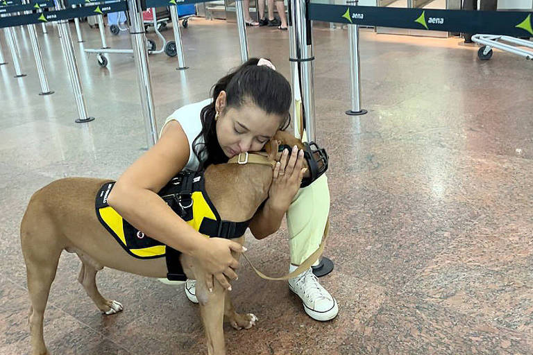 Uma mulher está agachada em um aeroporto， abraçando um cachorro de raça mista que usa um colete amarelo. O ambiente ao fundo mostra filas de check-in e pessoas se movendo. A mulher parece feliz e carinhosa com o animal.