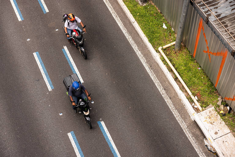 A imagem mostra uma vista aérea de uma avenida com duas motocicletas. Um motociclista está usando um capacete azul e uma mochila， enquanto o outro está vestido com uma camiseta clara e um capacete. As faixas da avenida são demarcadas por linhas azuis. À direita da imagem， há uma cerca de metal e um espaço verde.