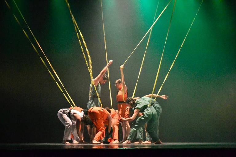 A imagem mostra um grupo de dançarinos em uma apresentação no palco. Eles estão usando roupas de cores variadas, como laranja, verde e cinza. Os dançarinos estão interagindo entre si, com alguns segurando fios amarelos que se estendem do teto, criando uma composição dinâmica. A iluminação é suave, com um fundo verde, que destaca os movimentos dos dançarinos.