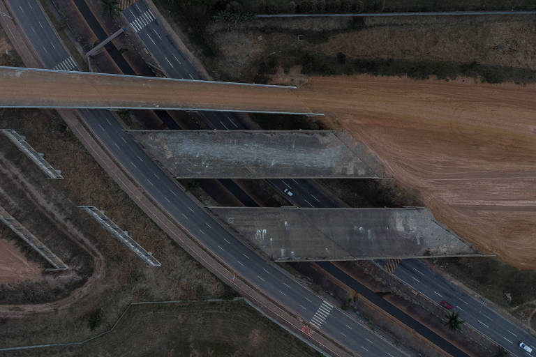 A imagem mostra uma vista aérea de uma rodovia em construção. Há seções de asfalto e concreto visíveis， com áreas de terra exposta ao redor. Algumas faixas da rodovia estão inacabadas， e há uma interseção visível. A vegetação ao redor é escassa， e a cena é predominantemente em tons de cinza e marrom.