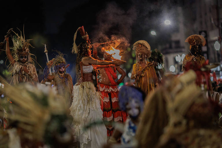 A imagem mostra um grupo de pessoas em trajes tradicionais, participando de uma celebração noturna. Algumas pessoas estão usando máscaras e adornos feitos de materiais naturais, como folhas e penas. Uma mulher no centro está segurando uma tocha acesa, enquanto outras dançam ao seu redor. O ambiente é iluminado por luzes ao fundo, criando uma atmosfera vibrante e festiva.