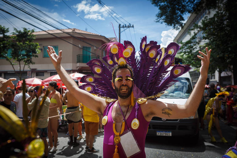 Um homem com um traje colorido e penas de pavão, levantando os braços em celebração durante um desfile de carnaval. Ao fundo, há uma multidão e veículos, com árvores e prédios ao redor.