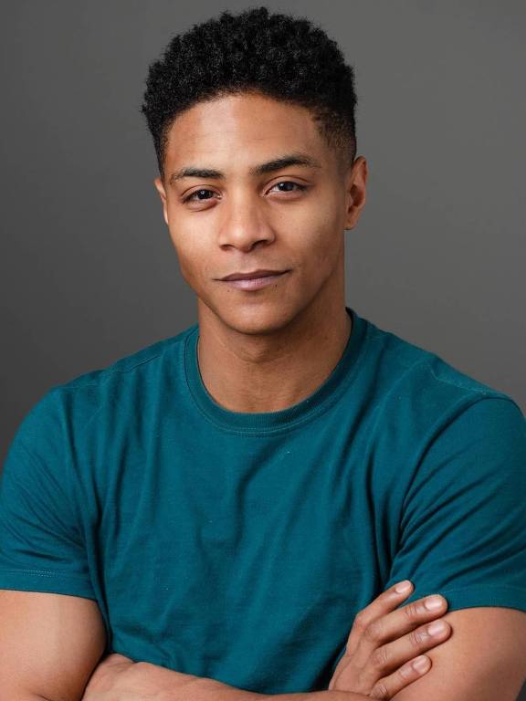 Um homem jovem está posando para a foto com os braços cruzados. Ele usa uma camiseta azul-escura e tem cabelo curto e encaracolado. O fundo é de uma cor cinza suave, criando um contraste com sua camiseta.