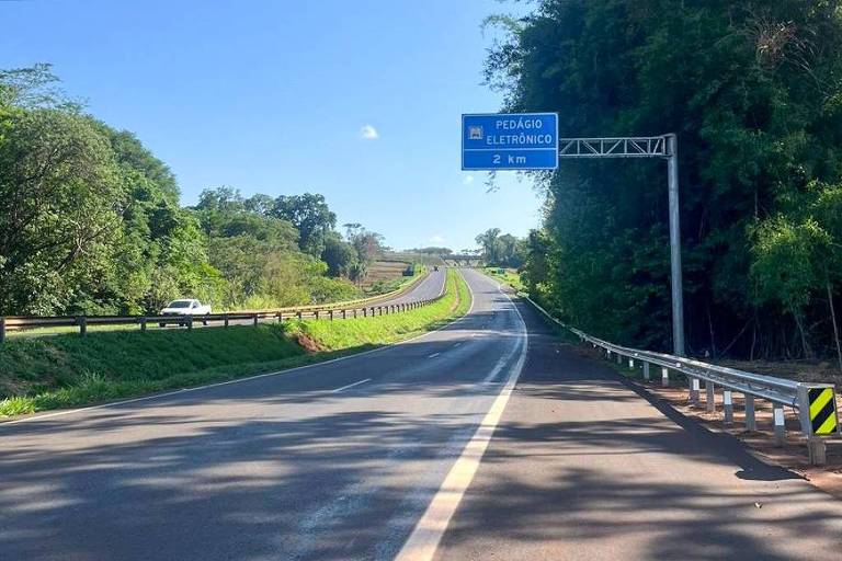 Imagem de uma estrada com vegetação ao lado. Há um sinal azul visível à frente， indicando direções. O céu está claro e ensolarado， e a estrada é pavimentada com uma faixa central branca. À direita， há uma barreira de proteção