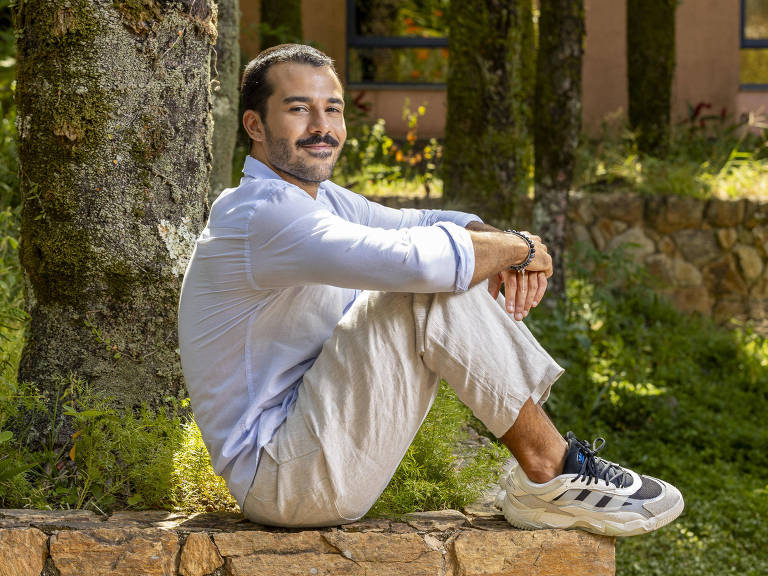 Um homem está sentado em uma pedra em um ambiente natural, cercado por árvores e vegetação. Ele usa uma camisa de manga longa clara e calças claras. O homem tem um bigode e está sorrindo, com as pernas cruzadas e as mãos apoiadas nos joelhos.