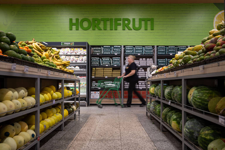 A imagem mostra setor de hortifrúti em um supermercado， com prateleiras repletas de frutas e verduras. No fundo， há uma parede verde com a palavra quot;hortifrutiquot; em letras grandes. À esquerda， há bananas e melões， enquanto à direita há melancias e outros produtos. Uma pessoa está passando com um carrinho de compras.