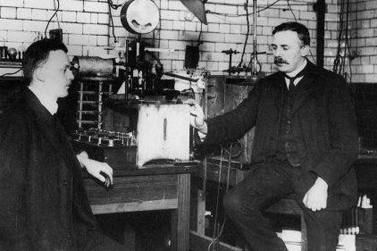 A imagem mostra dois homens em um laboratório. Um deles está sentado à esquerda， enquanto o outro está à direita， segurando um dispositivo. O ambiente é composto por mesas de trabalho e equipamentos científicos ao fundo， com paredes de tijolos e fios pendurados.