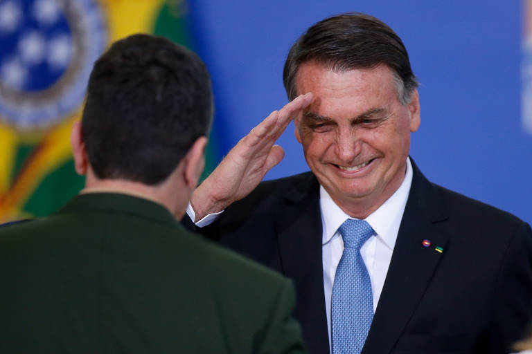 A imagem mostra um homem sorridente, com cabelo escuro e terno, fazendo um gesto de saudação. Ele está em um evento formal, com um fundo que inclui a bandeira do Brasil. À sua frente, um homem de uniforme militar está de costas, com a mão levantada em direção ao homem que saúda.