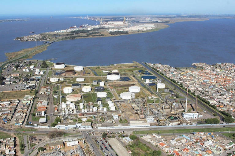 A imagem mostra uma vista aérea de uma área industrial， localizada próxima a um grande corpo de água. Na imagem， há diversos tanques de armazenamento grandes e estruturas industriais， possivelmente pertencentes a uma refinaria ou planta petroquímica. À direita， vê-se uma área residencial com casas agrupadas. Ao fundo， há mais instalações industriais à beira do corpo de água. A cena é ampla e captura tanto a área urbana quanto a zona industrial adjacente à água.

