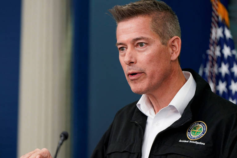 Sean Duffy é um homem branco vestindo um paletó preto com um emblema de investigação de acidentes fala em uma conferência de imprensa. Ele está gesticulando com a mão direita. Ao fundo， há uma parede azul e uma bandeira dos Estados Unidos.