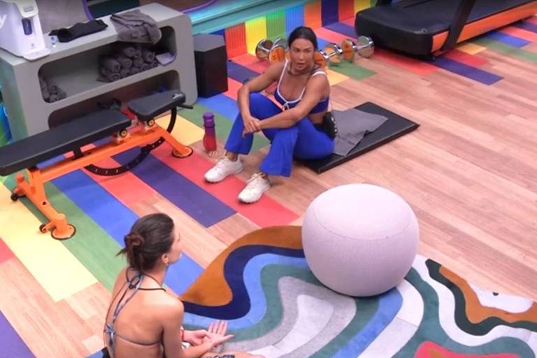 A imagem mostra duas pessoas em um ambiente de treino. Uma delas está sentada no chão, de costas, com cabelo preso e usando um biquíni. A outra pessoa está sentada em um banco, vestindo uma blusa sem mangas e calças azuis, com uma garrafa de água ao lado. No fundo, há equipamentos de treino, como um banco e pesos, e o chão é colorido com um padrão de listras. Um grande objeto redondo e claro está no centro da cena.