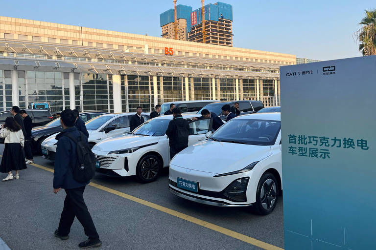 A imagem mostra uma exibição de carros elétricos em um evento ao ar livre. Vários veículos estão alinhados em uma área pavimentada， com pessoas interagindo ao redor deles. Ao fundo， há um edifício com janelas grandes e em construção， além de um painel informativo à direita， que contém texto em chinês. O céu está claro e a iluminação é suave.