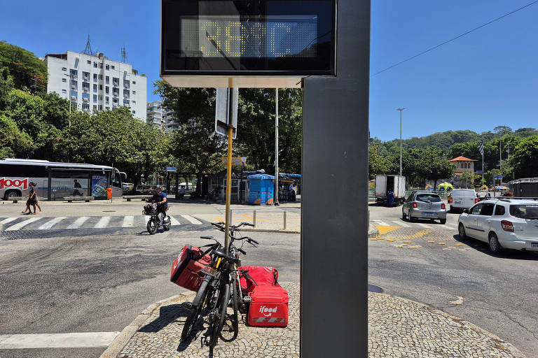 A imagem mostra um cruzamento urbano em um dia ensolarado. À esquerda， há um semáforo digital que indica a hora. Em primeiro plano， duas bicicletas estão estacionadas em um suporte， com bolsas vermelhas de entrega penduradas nelas. Ao fundo， é possível ver ônibus e carros em movimento， além de prédios e árvores ao redor.