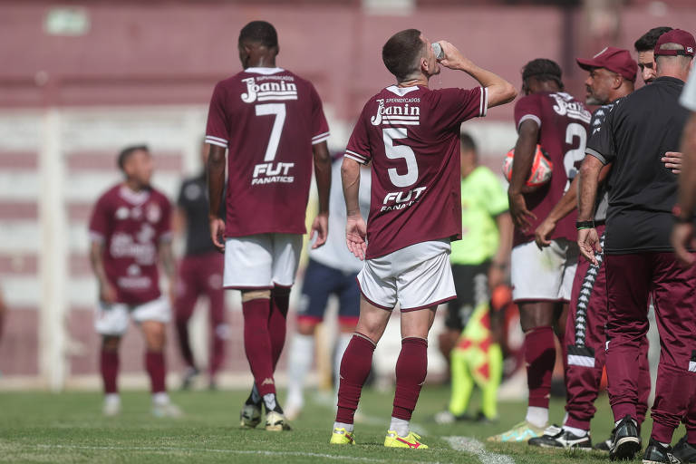 A imagem mostra jogadores de futebol em um campo. Um jogador está usando a camisa número 5, enquanto outro está com a camisa número 7. Ambos estão vestidos com uniformes marrom e branco. Há também um grupo de pessoas à beira do campo, incluindo um treinador. O ambiente parece ser um dia ensolarado.