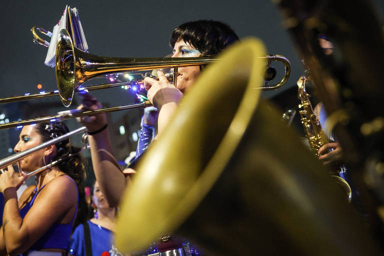 A imagem mostra uma banda de música em performance, com foco em instrumentos de metal, como trombones e uma tuba. Algumas pessoas estão visíveis, vestindo roupas coloridas e segurando seus instrumentos. O fundo é desfocado, mas há luzes e uma atmosfera festiva.