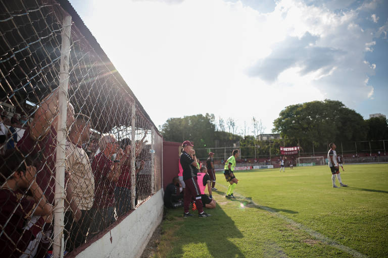 A imagem mostra um campo de futebol com uma torcida visível atrás de uma rede. À esquerda， há pessoas assistindo ao jogo， algumas vestindo camisetas de cor marrom. No campo， um jogador está se preparando para entrar em campo， enquanto outro jogador está em ação. O céu está parcialmente nublado， com luz do sol iluminando a cena.