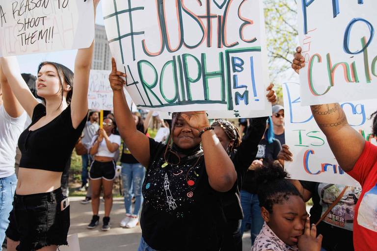 A imagem mostra um grupo de pessoas participando de um protesto. Uma mulher à esquerda levanta os braços， enquanto uma mulher no centro segura um cartaz que diz 039;JUSTICE Ralphie BLM039;. Ao lado dela， uma criança observa. O ambiente parece ser uma manifestação em apoio a uma causa social.