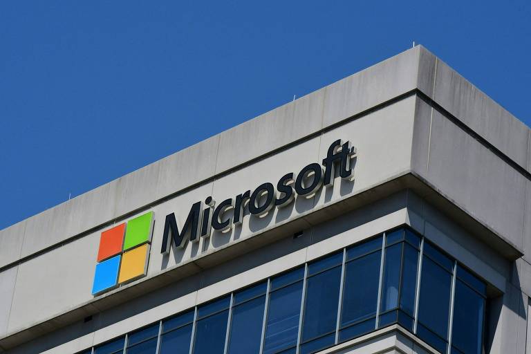 Imagem do topo de um edifício com o logotipo da Microsoft em destaque. O logotipo é composto por um texto em letras minúsculas e um ícone colorido com quatro quadrados (azul， verde， amarelo e vermelho) dispostos em um formato de janela. O céu está claro e azul ao fundo.