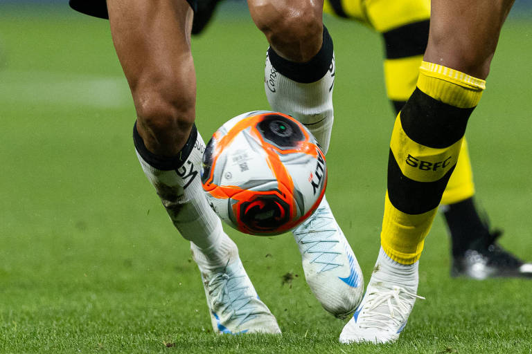 Dois jogadores de futebol estão em campo， disputando a bola. O jogador à esquerda veste uma camisa branca com detalhes pretos e o número 20， enquanto o jogador à direita usa uma camisa preta com detalhes amarelos. O fundo mostra um estádio com torcedores.