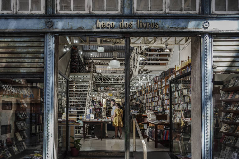 A imagem mostra a entrada de uma livraria chamada 039;Ler de Lado039;. A fachada é composta por uma porta de vidro e uma estrutura metálica. Dentro da loja， é possível ver prateleiras repletas de livros e uma escada ao fundo. Há uma pessoa em pé perto do balcão de atendimento， e a iluminação é suave， criando um ambiente acolhedor.