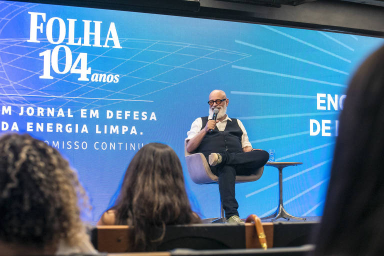 A imagem mostra um evento da Folha de S.Paulo， com um homem de óculos e barba sentado em uma cadeira， falando ao microfone. Ao fundo， há um painel azul com o texto 039;FOLHA 104 anos039; e 039;UM JORNAL EM DEFESA DA ENERGIA LIMPA. COMPROMISSO CONTÍNUO.039; A plateia é visível em primeiro plano， com pessoas de costas.