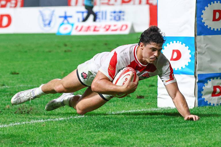 Um jogador de rugby está se estendendo para marcar um try， com a bola na mão. Ele está vestindo um uniforme branco com detalhes vermelhos e pretos. O campo é verde e há uma marcação de try ao fundo. O jogador parece estar em movimento rápido， com o corpo inclinado para frente.