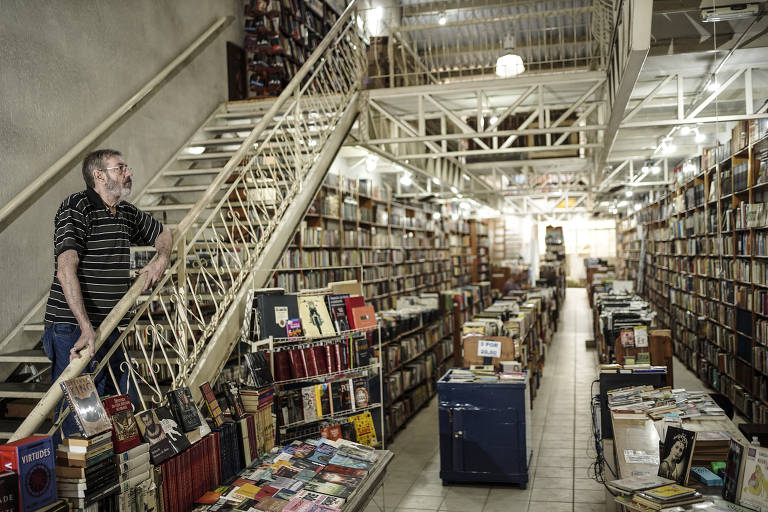 A imagem mostra o interior de uma livraria ampla e bem iluminada, com estantes repletas de livros. À esquerda, uma escada de metal leva a um andar superior. Um homem de cabelos grisalhos e barba está em pé, apoiado na grade da escada, observando o ambiente. O chão é de cerâmica clara e há mesas com livros expostos em várias partes do espaço.