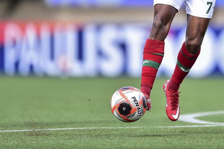 A imagem mostra a perna de um jogador de futebol em movimento， prestes a chutar uma bola. O jogador está usando uma camisa branca com detalhes em verde e vermelho， e meias vermelhas. A bola é de futebol， com a inscrição 039;PENALTY039; visível. Ao fundo， há uma faixa com a palavra 039;PAULISTÃO039;.