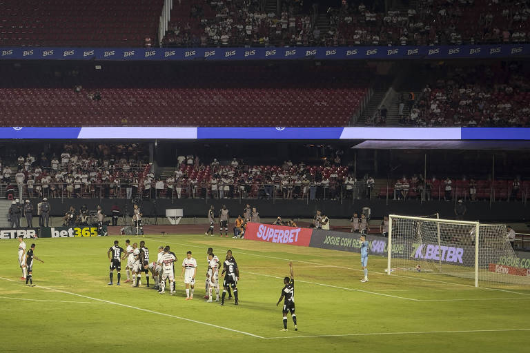 A imagem mostra um campo de futebol em um estádio， onde jogadores de duas equipes estão posicionados para uma cobrança de falta. Um goleiro está preparado na sua área， enquanto os jogadores adversários formam uma barreira. Ao fundo， há uma arquibancada com torcedores， e uma tela eletrônica visível na parte superior do estádio.