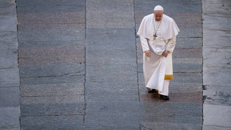 A imagem mostra um homem vestido com uma vestimenta branca， que parece ser um papa， caminhando em um espaço pavimentado. Ele está ajustando a parte inferior de sua vestimenta enquanto se move. O chão é composto por pedras em tons de cinza e há uma iluminação suave.