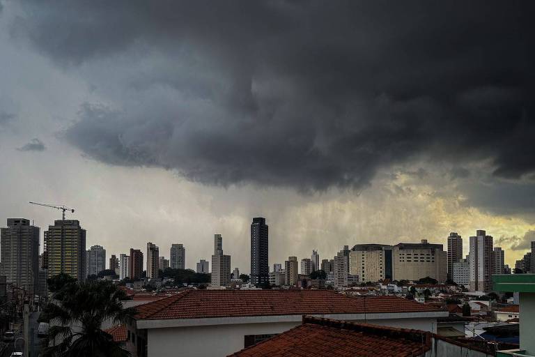 A imagem mostra uma vista urbana com prédios altos sob um céu nublado e escuro. As nuvens são densas e ameaçadoras， e há uma leve iluminação ao fundo， sugerindo que pode estar chovendo. O primeiro plano apresenta telhados de casas， com algumas árvores visíveis.