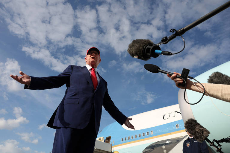 Trump， de terno escuro e gravata vermelha， está em pé com os braços abertos em frente a um avião. O céu está parcialmente nublado. Microfones estão apontados para ele.