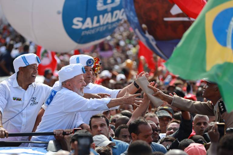 A imagem mostra um grande grupo de pessoas em um evento político ao ar livre. No centro， três homens， um deles vestido com uma camisa branca e chapéu， estão estendendo as mãos para o público. O homem à esquerda também usa uma camisa branca， enquanto o da direita está com uma camisa escura. Ao fundo， há bandeiras vermelhas e uma bandeira verde e amarela do Brasil. Balões e uma grande faixa com a palavra 039;SALVADOR039; são visíveis.