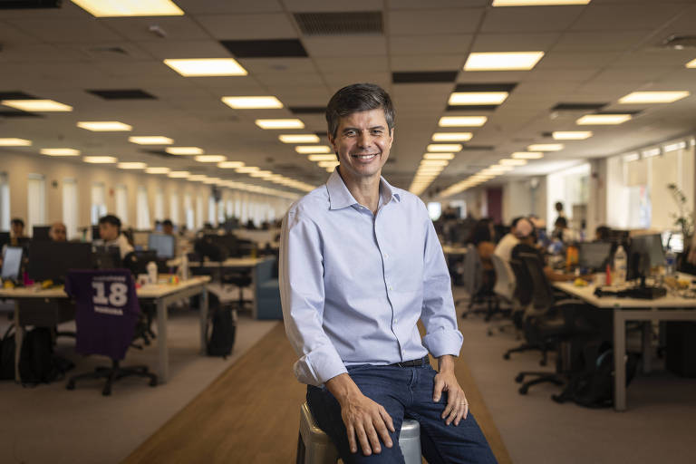 Um homem sorridente está sentado em um banco em um escritório moderno. Ele usa uma camisa de botão clara e jeans. Ao fundo， há várias mesas de trabalho ocupadas por pessoas， com computadores e materiais de escritório visíveis. A iluminação é clara e o ambiente parece bem organizado.