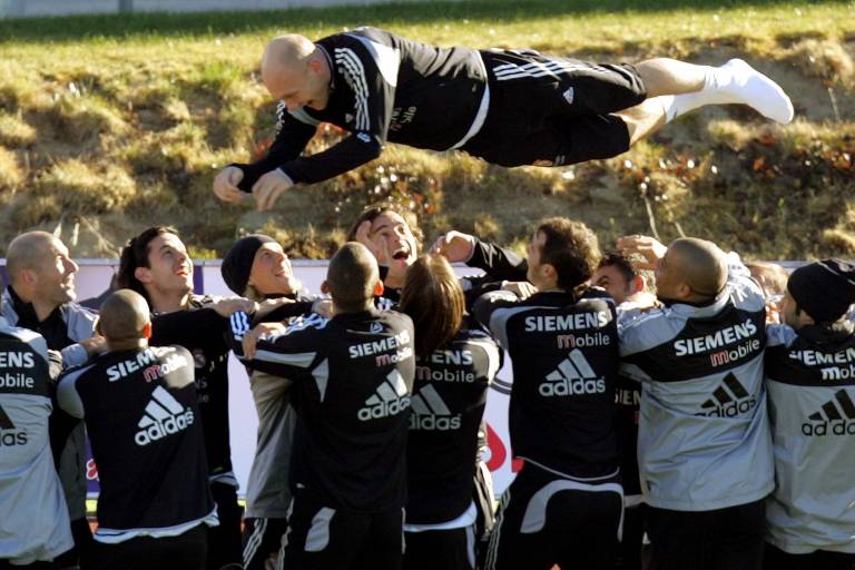 Um grupo de jogadores de futebol está em uma celebração， com um jogador sendo levantado e lançado no ar por seus companheiros. Eles estão vestidos com uniformes de treino， predominantemente em preto e cinza， com logotipos da Siemens e Adidas visíveis. O ambiente parece ser um campo de treinamento， com grama ao fundo e uma atmosfera de alegria e camaradagem.