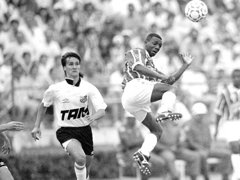 A imagem mostra um momento de um jogo de futebol em preto e branco. Um jogador， vestido com uma camisa listrada， está saltando para chutar a bola que está no ar. Outro jogador， usando uma camisa branca com a palavra 039;TAM039; em destaque， está correndo em direção ao jogador que está chutando. O fundo é composto por uma multidão de torcedores.