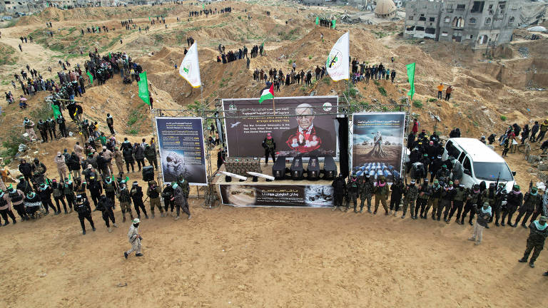 Imagem mostra a entrega dos corpos dos reféns em uma área desértica， com um grande grupo de pessoas reunidas em um terreno arenoso. Há bandeiras verdes do Hamas e uma estrutura montada com painéis que exibem imagens e textos acima de um palco， onde há quatro caixões. Os combatentes da facção estão vestidos com uniformes escuros， formando uma linha em frente aos painéis. Ao fundo， há edifícios em ruínas e uma van branca estacionada.