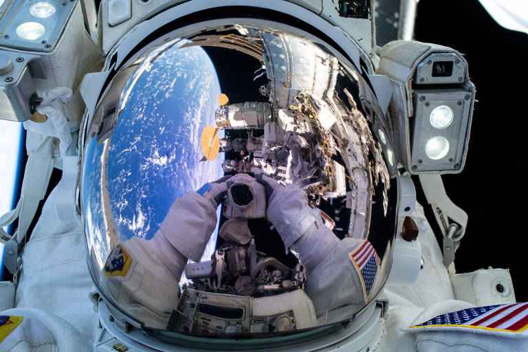 A imagem mostra um astronauta em um traje espacial， com um visor refletindo a Terra e a Estação Espacial Internacional. O reflexo no visor exibe a superfície do planeta e parte da estrutura da estação. O traje do astronauta possui emblemas e luzes， e o fundo é predominantemente negro， com a Terra visível em azul e branco.