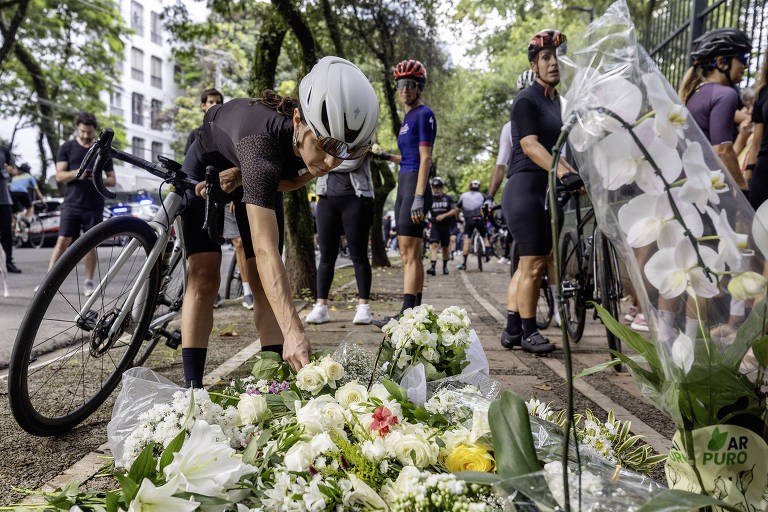 A imagem mostra um grupo de ciclistas em uma homenagem. Um ciclista， usando um capacete e roupas de ciclismo， está se agachando para colocar flores brancas em um monte de flores que está no chão. Ao fundo， outros ciclistas estão parados， alguns em bicicletas， e há árvores e prédios ao redor. A cena transmite um sentimento de respeito e solidariedade.