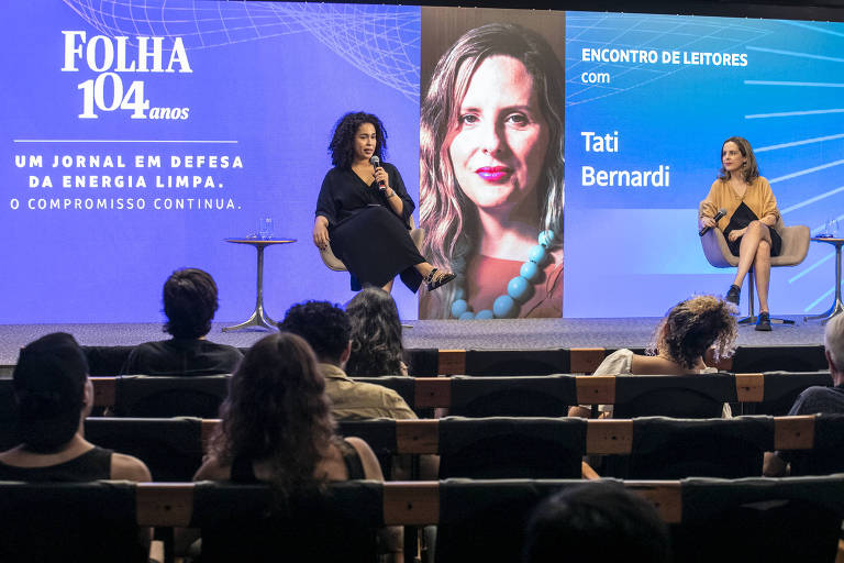 A imagem mostra um evento chamado 'Encontro de Leitores' com Tati Bernardi. No fundo, há um painel azul com o texto 'ENCONTRO DE LEITORES com Tati Bernardi'. À esquerda, uma mulher sentada em uma cadeira, segurando um microfone, e à direita, outra mulher sentada em uma cadeira, olhando para a primeira. O fundo também exibe o logotipo da Folha, com a frase 'UM JORNAL EM DEFESA DA ENERGIA LIMPA. O COMPROMISSO CONTINUA.'