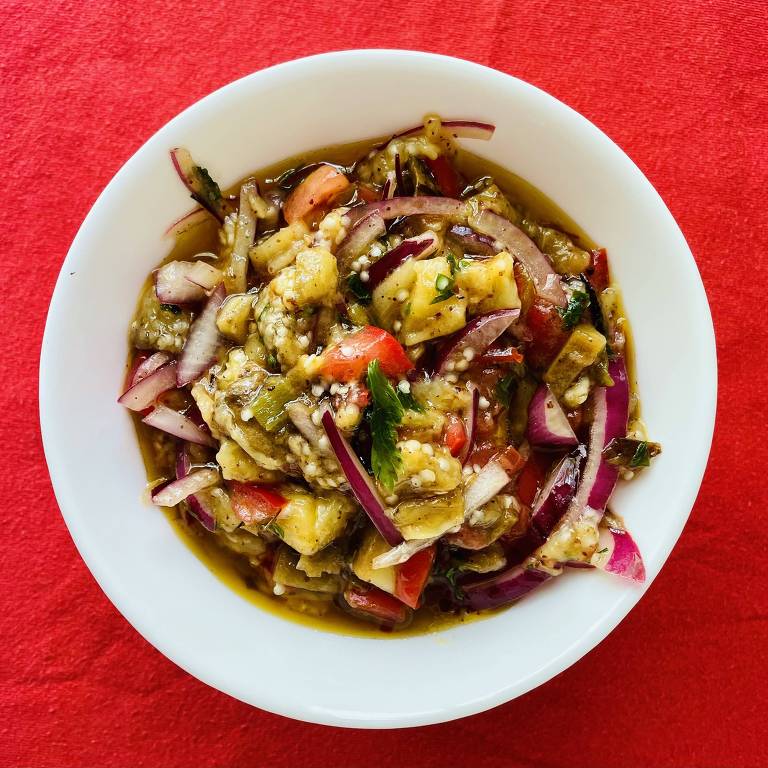 Uma salada servida em uma tigela branca， contendo pedaços de vegetais como tomate， cebola roxa， pimentão e ervas， misturados em um molho. O fundo da imagem é vermelho.