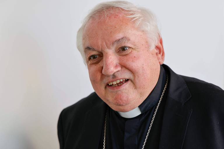 Um homem idoso, com cabelo grisalho e pele clara, está sorrindo. Ele usa uma batina preta e um colarinho clerical branco. O fundo é claro e neutro.