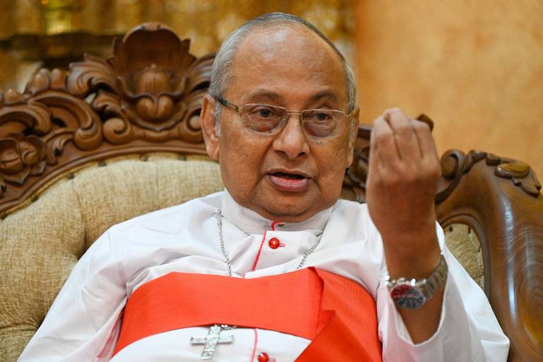 Um cardeal está sentado em uma cadeira ornamentada, vestindo uma batina branca com um cinto vermelho. Ele está gesticulando com a mão direita, enquanto a esquerda repousa sobre o braço da cadeira. O fundo é decorado com elementos dourados.